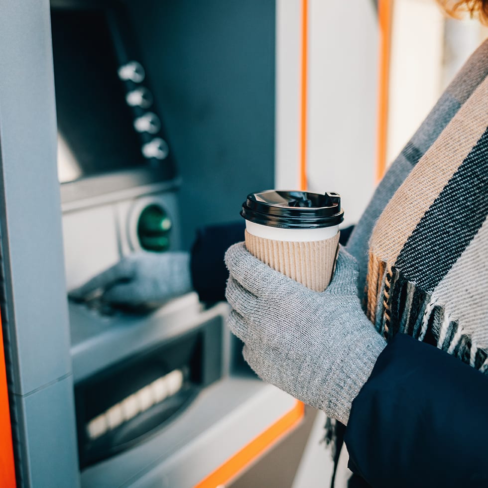Close-up of young woman in warm clothing holding coffee and using an ATM on city street in winter day.