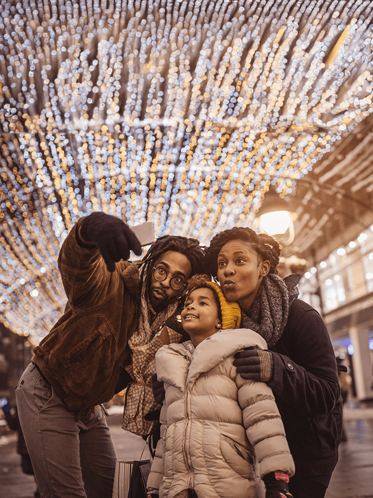 A family of three is taking a selfie while in a tunnel that is festive and decorated with lots of holiday lights